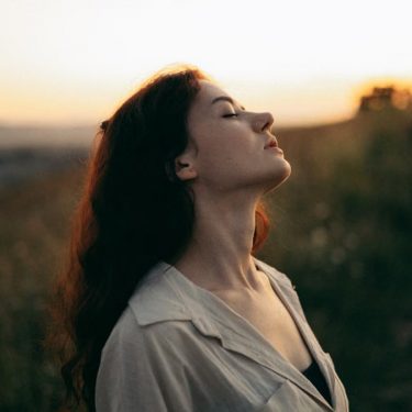 Woman standing with eyes closed in a field during sunset, exuding tranquility and peace.